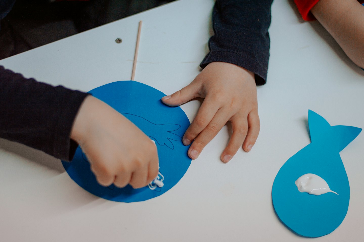 Children glue paper craft pieces together on a white table.