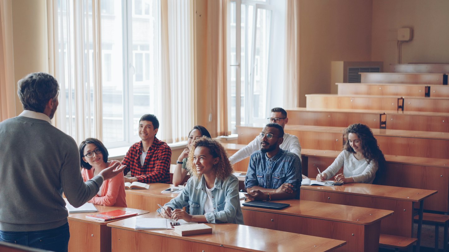 Teacher addresses diverse students in a brightly lit lecture hall.
